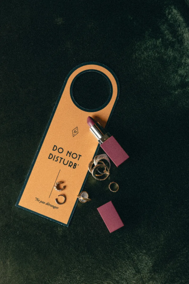 Une pancarte de porte orange avec l'inscription « Do Not Disturb » et « Ne pas déranger », accompagnée d'un rouge à lèvres violet et de plusieurs bagues dorées sur un fond sombre.