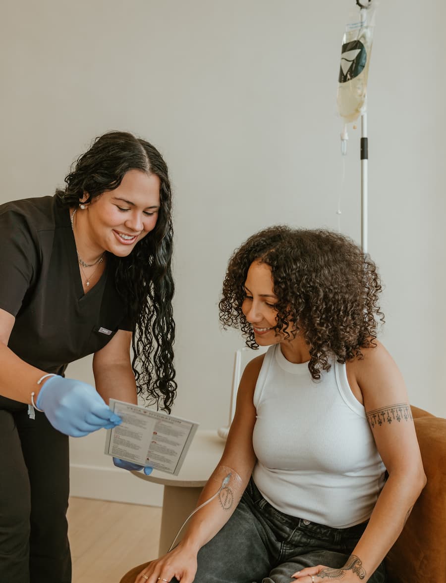 Healthcare professional in black scrubs with gloves showing information to a seated patient receiving IV treatment.