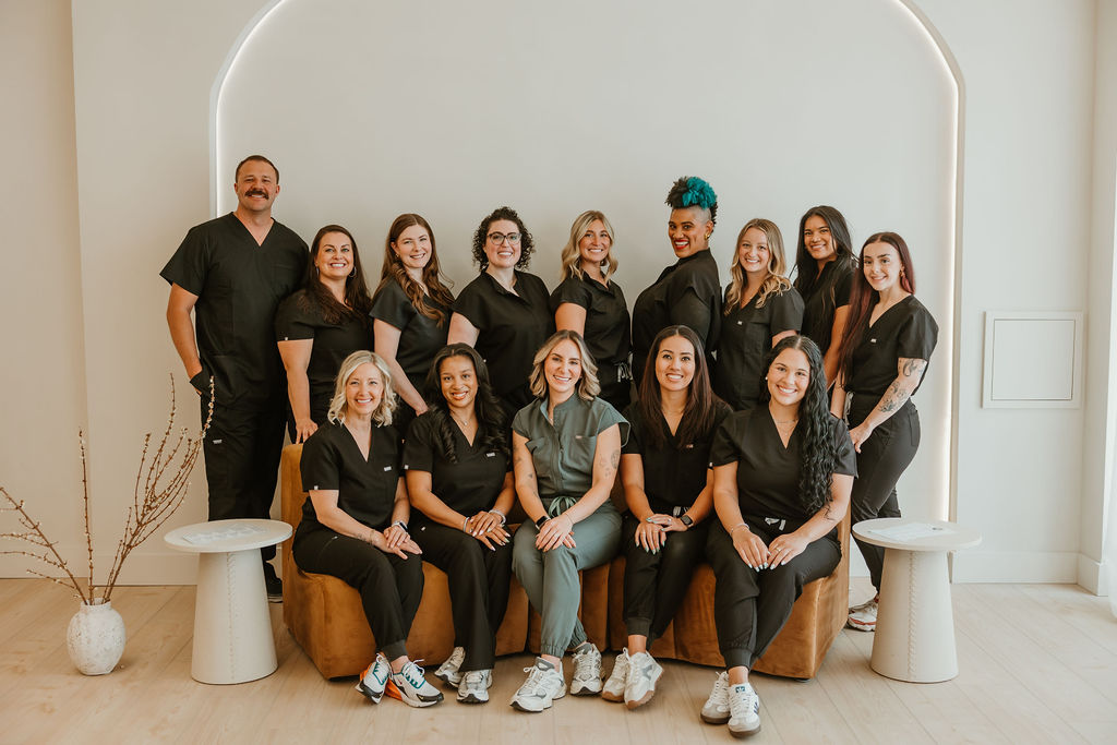 Group portrait of fourteen healthcare professionals in black scrubs, posing indoors with a neutral background and two small round tables on each side.