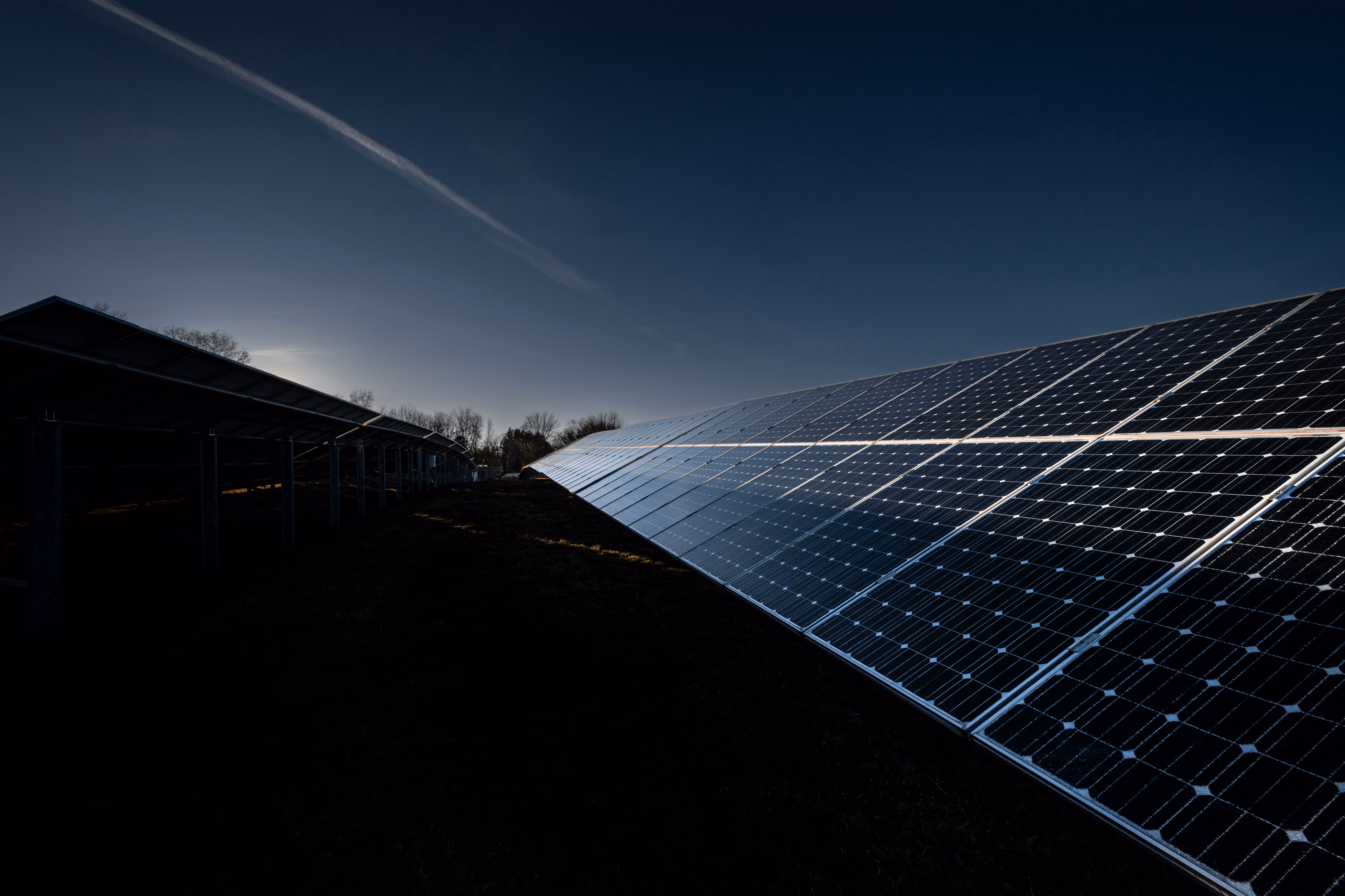 Rows of solar panels angled under a darkening sky at dusk with a faint contrail above.