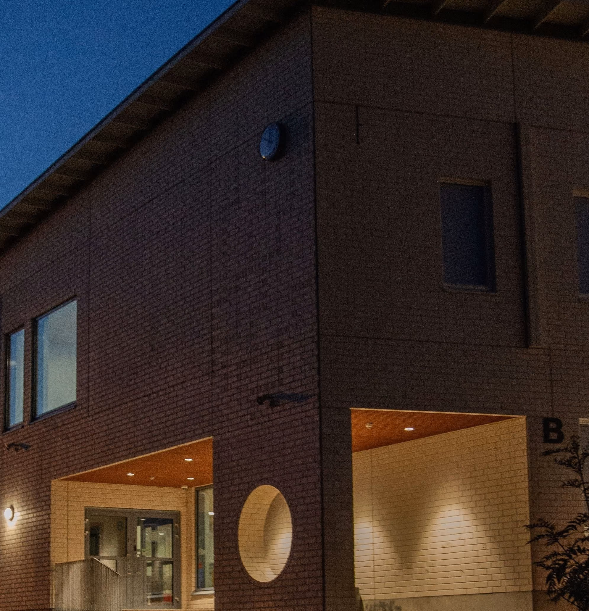 Modern brick building corner with a circular window and lit entrance area at night.