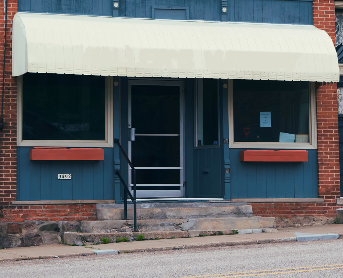 Closed storefront with beige awning, blue facade, two windows, and concrete steps with railing.