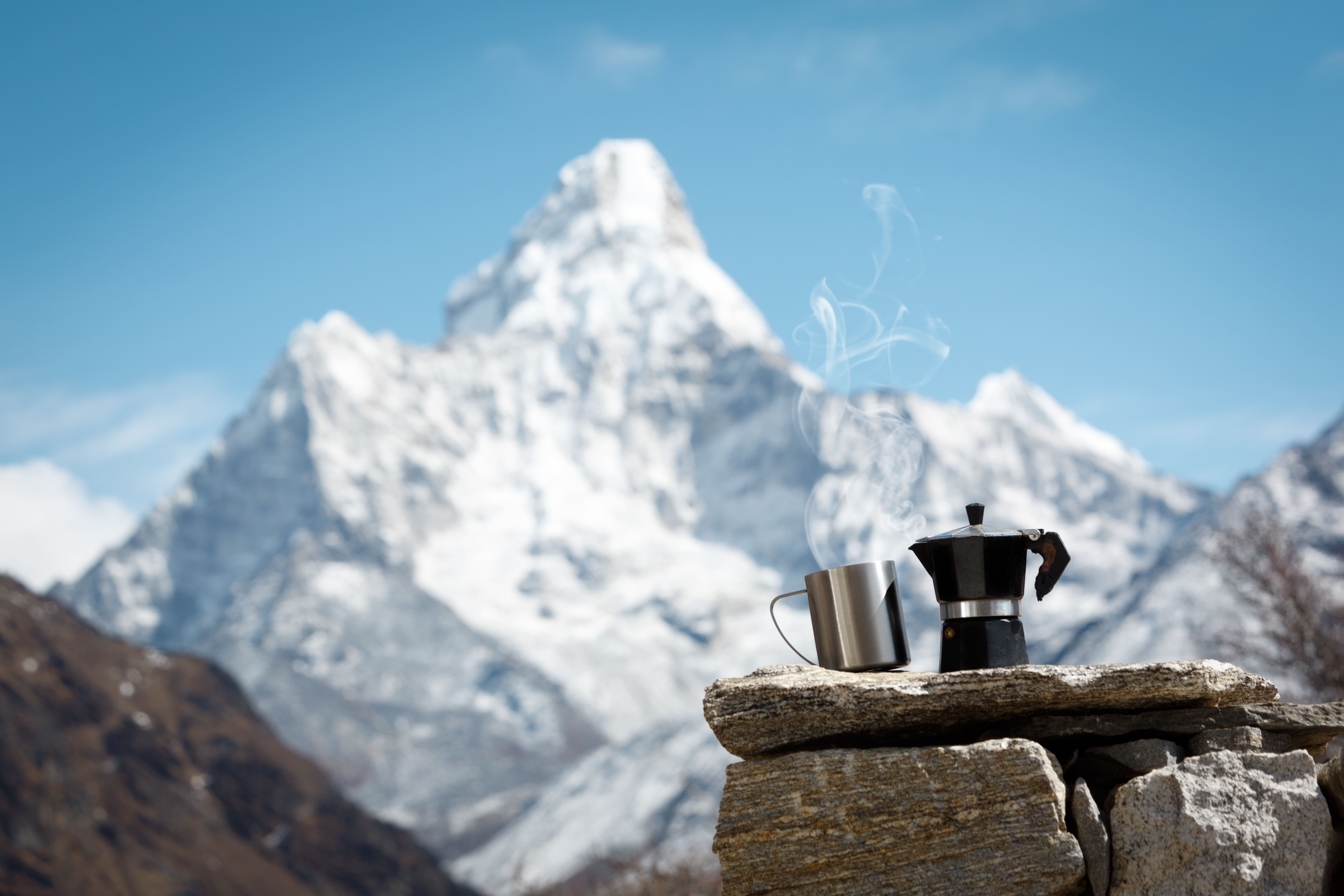 A stainless steel mug with a coffee maker on top of a pile of stones, behind is a view of glacier mountains
