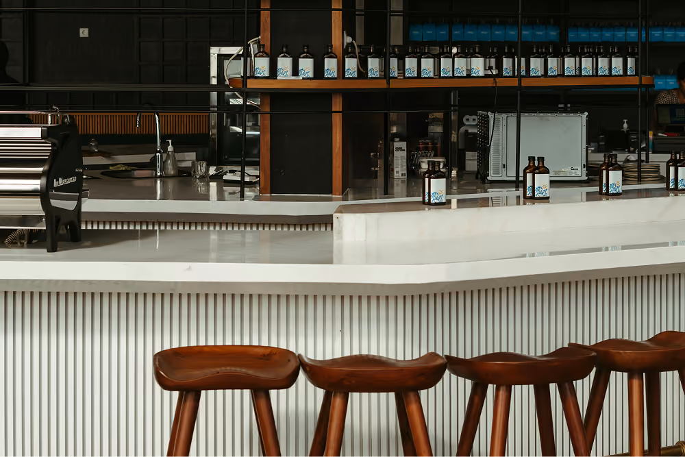Modern cafe counter with white surfaces, wooden stools, and rows of bottled products on shelves.