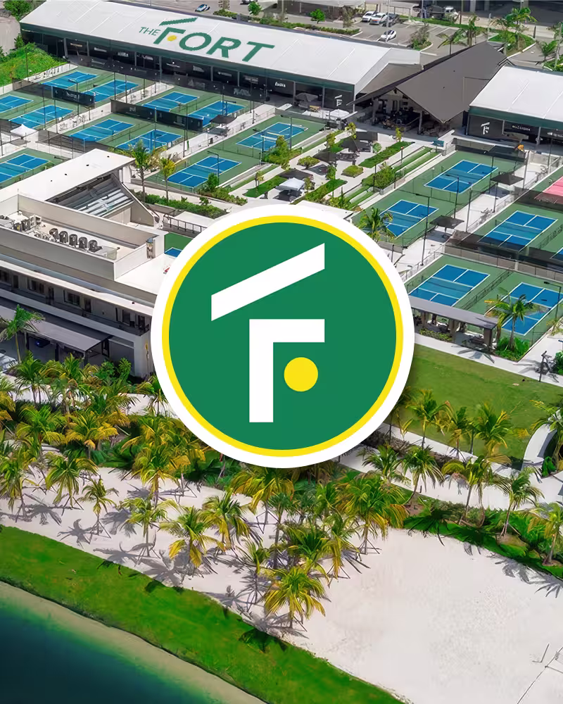 Aerial view of The Fort tennis complex with multiple courts, buildings, palm trees, and sand area.