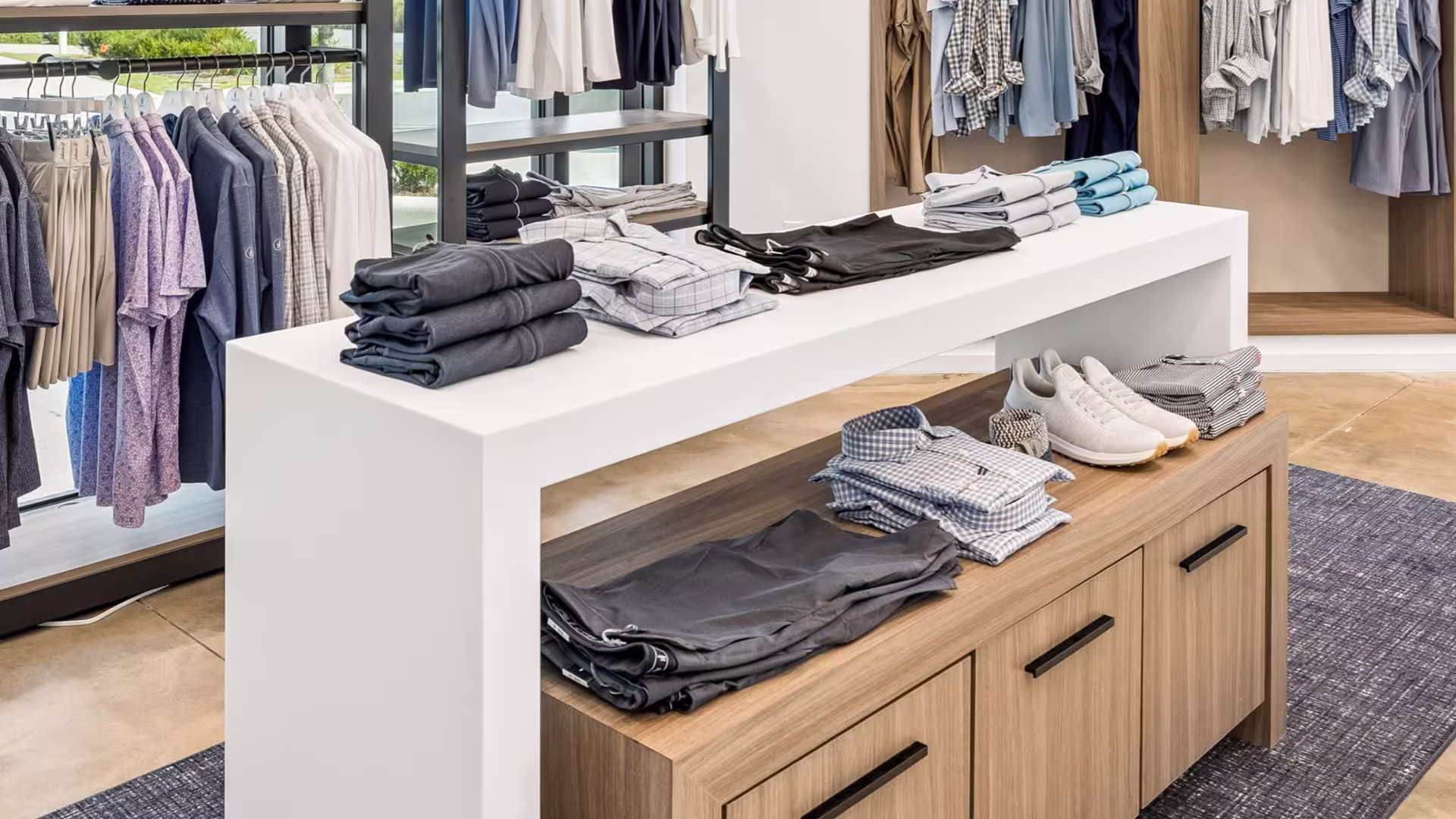 Clothing store display with folded shirts, pants, and white sneakers on white and wooden tables.