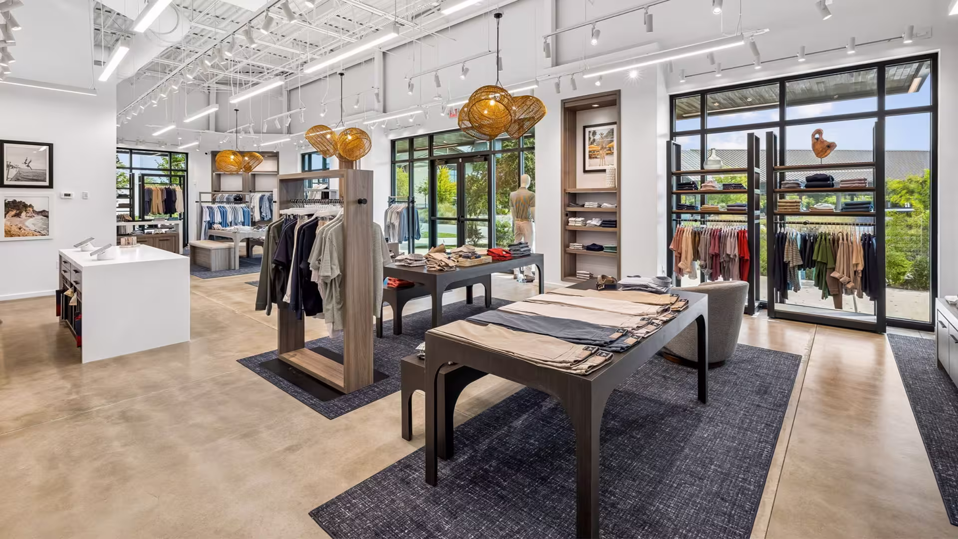 Modern clothing store interior with tables, racks of shirts, pants, and large windows letting in natural light.