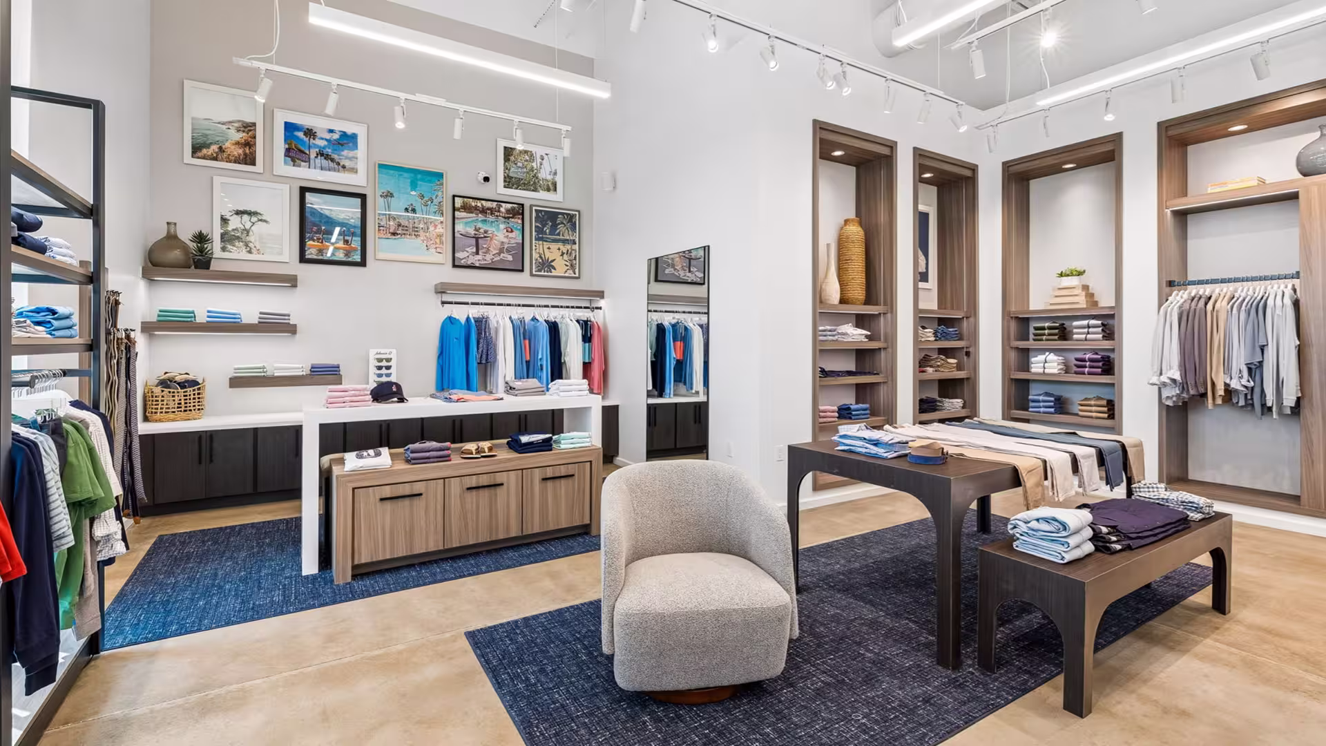 Modern clothing store interior with folded clothes, hanging shirts, blue rugs, and a gray armchair.