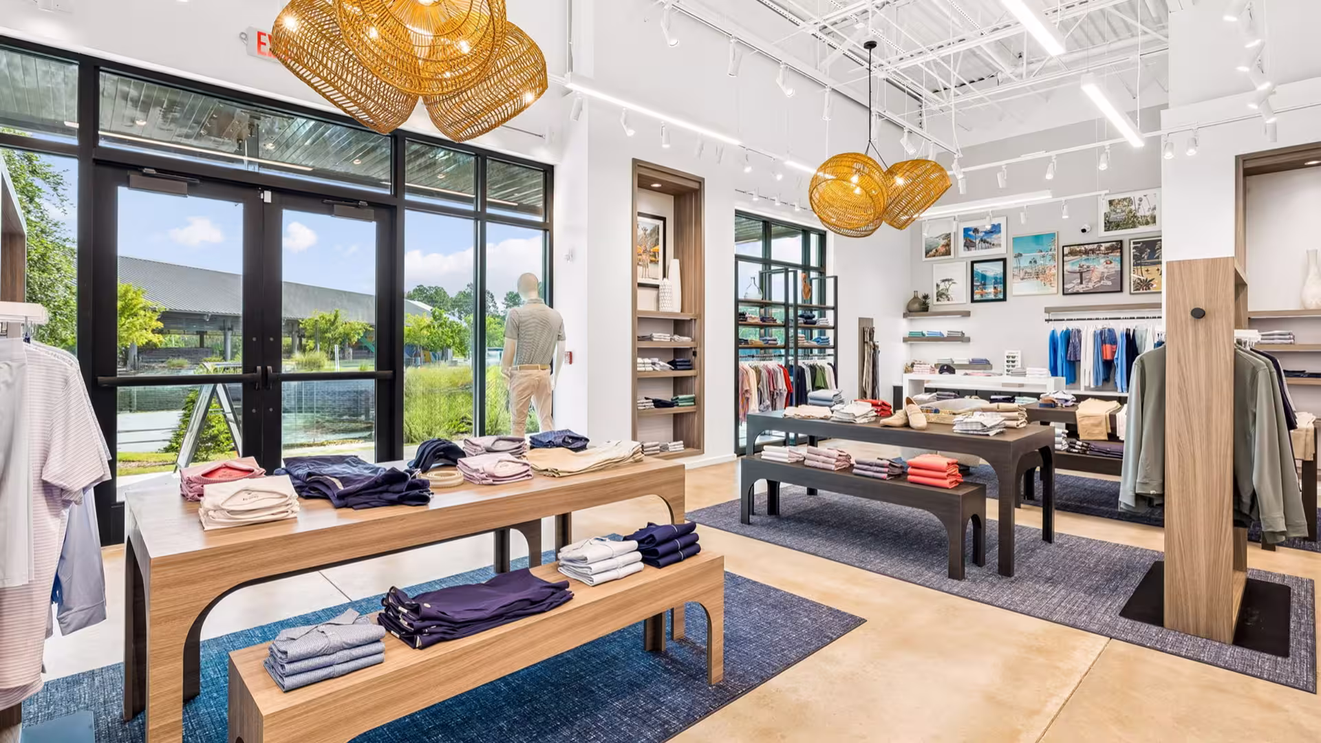 Bright clothing store interior with wooden tables, folded clothes, hanging racks, and wicker light fixtures.