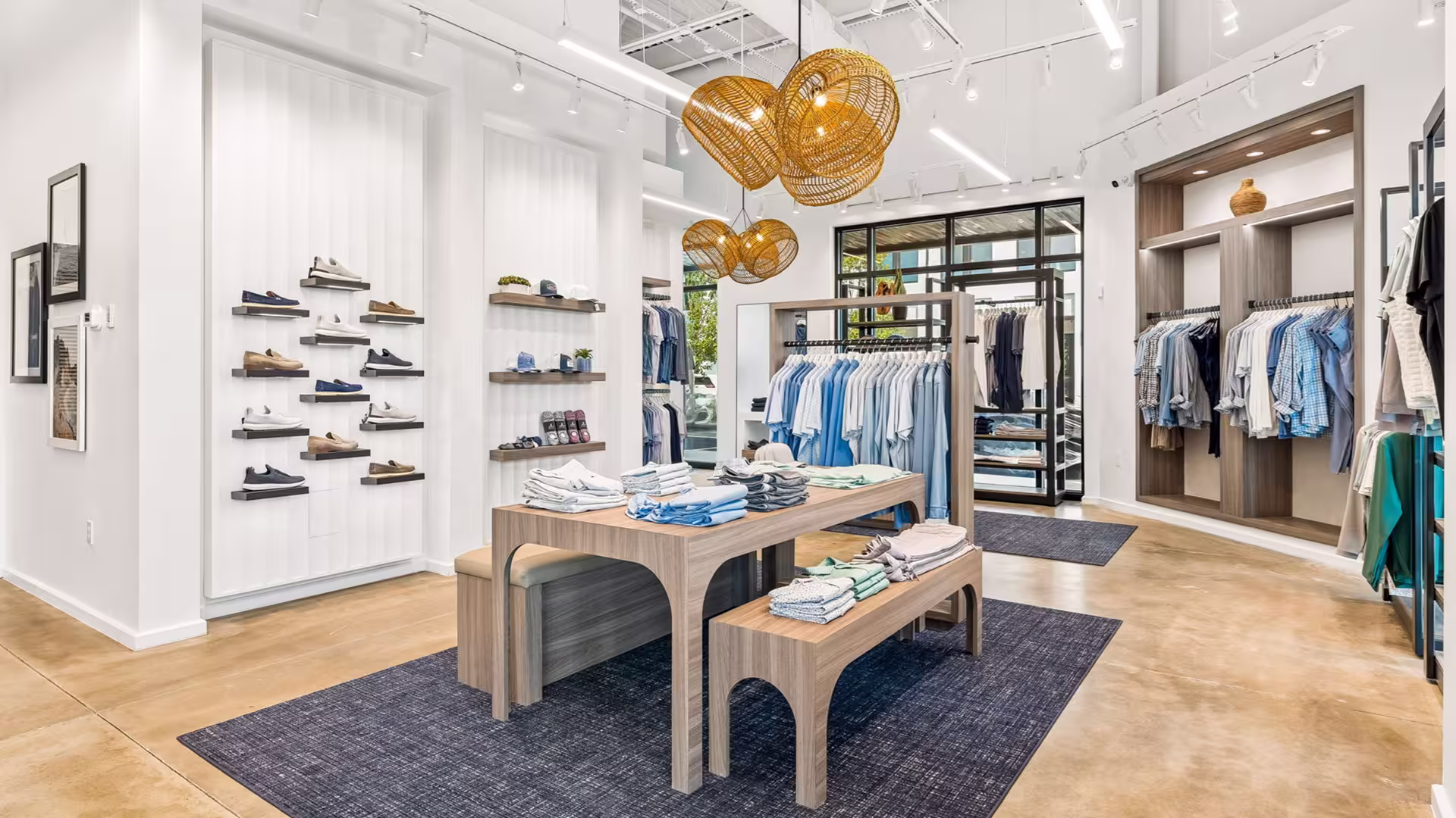 Bright modern clothing store with wooden table, benches, hanging lights, and apparel on racks and shelves.