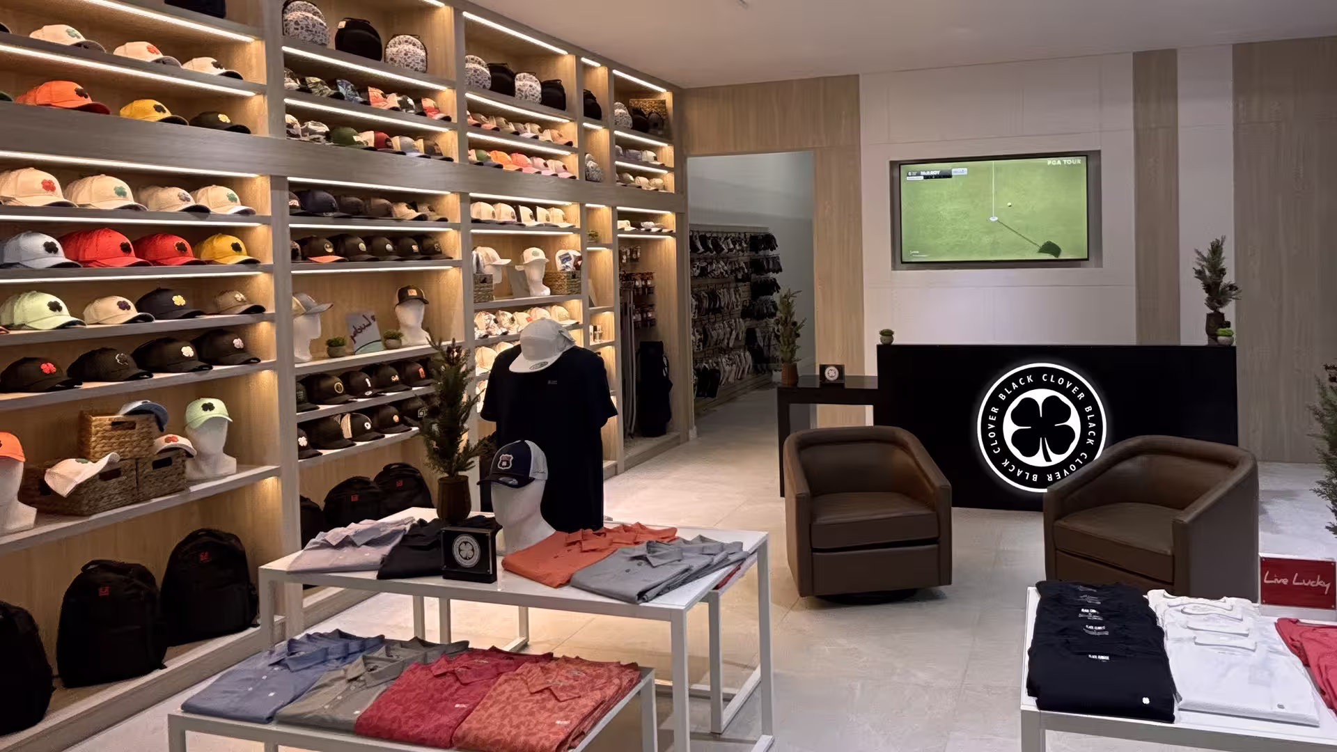 Retail store interior with shelves of hats, folded shirts, mannequins, chairs, and a TV showing a golf game.
