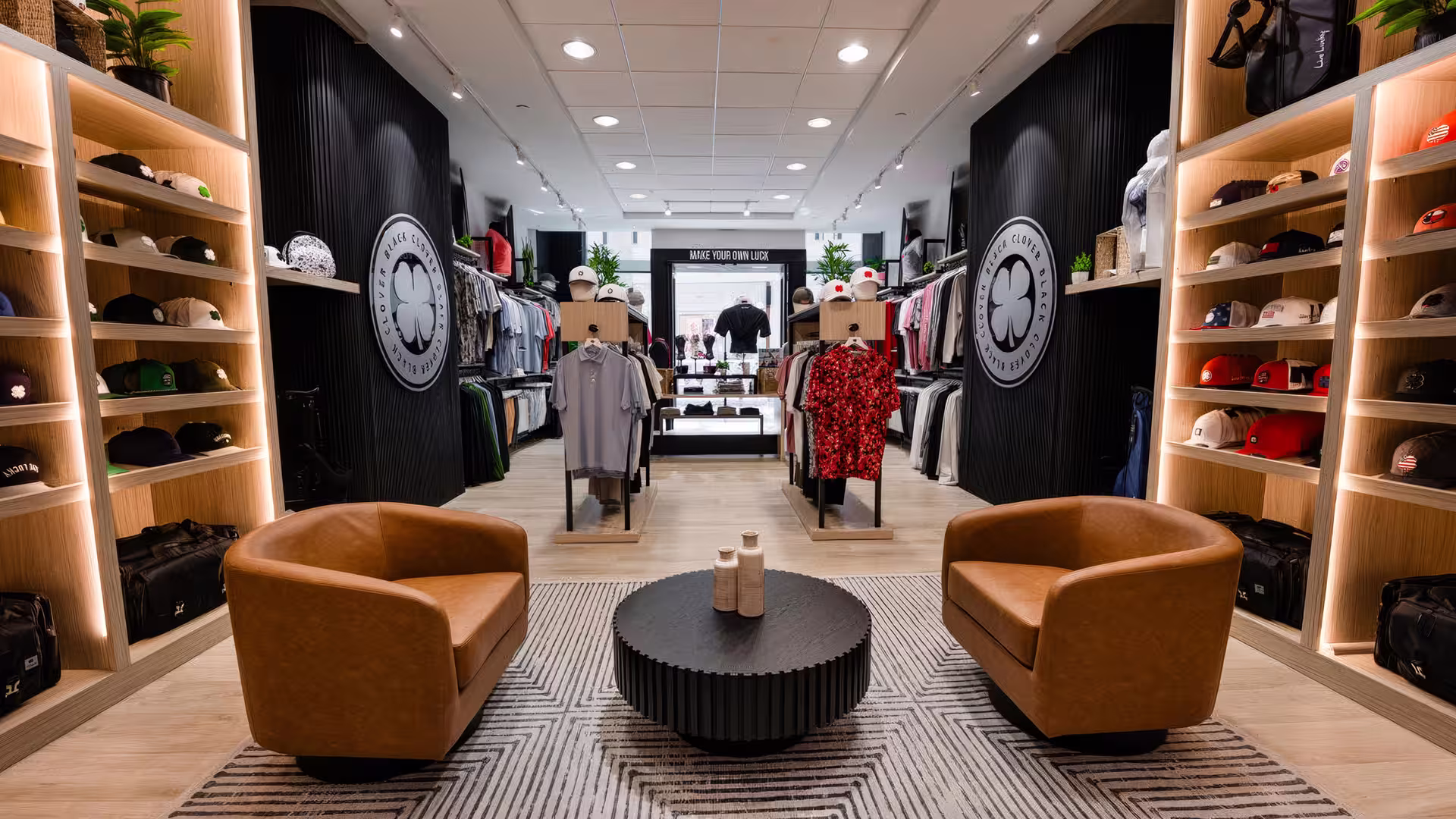 Modern retail store interior with leather chairs, a round table, hats, and clothing racks.