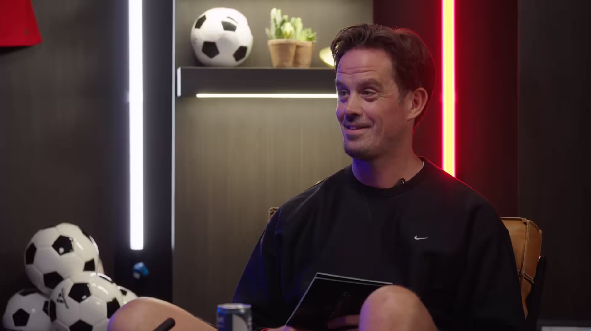 Smiling man in black Nike sweatshirt sitting with soccer balls and a can nearby.