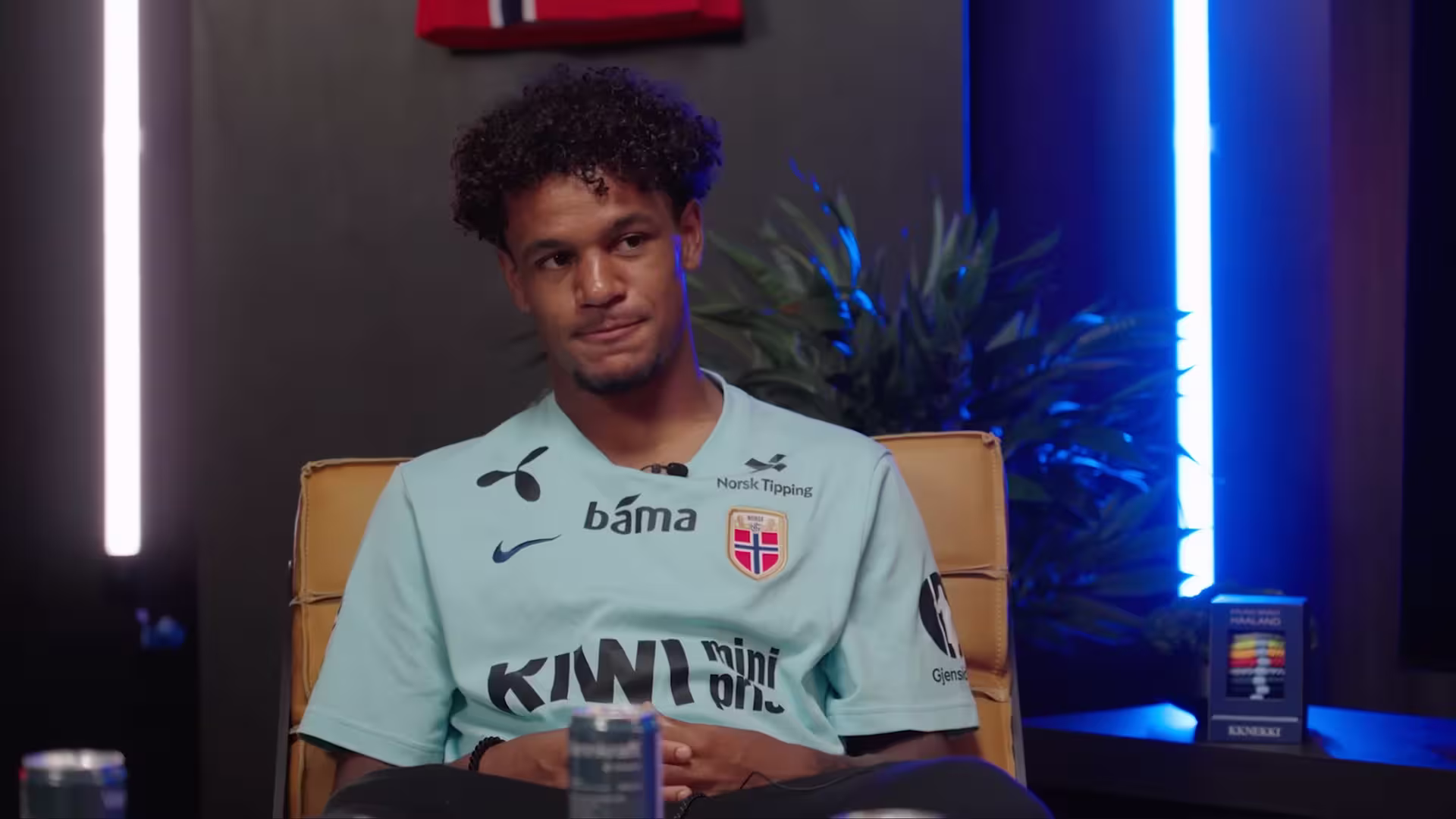 Young man with curly hair wearing a light blue Norway football jersey sitting on a chair.