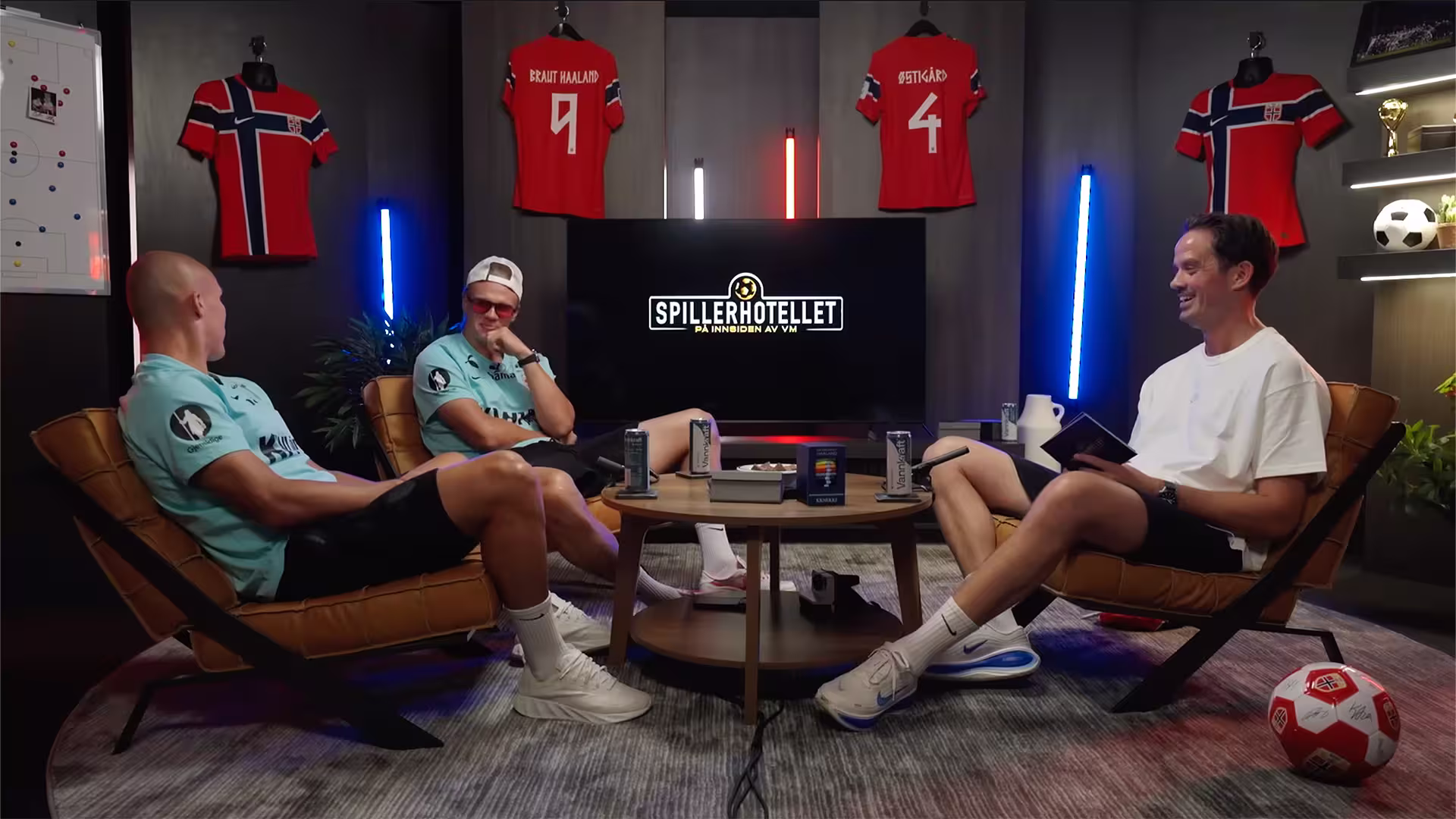 Three men sitting in chairs around a table in a room with Norwegian soccer jerseys and a TV screen.