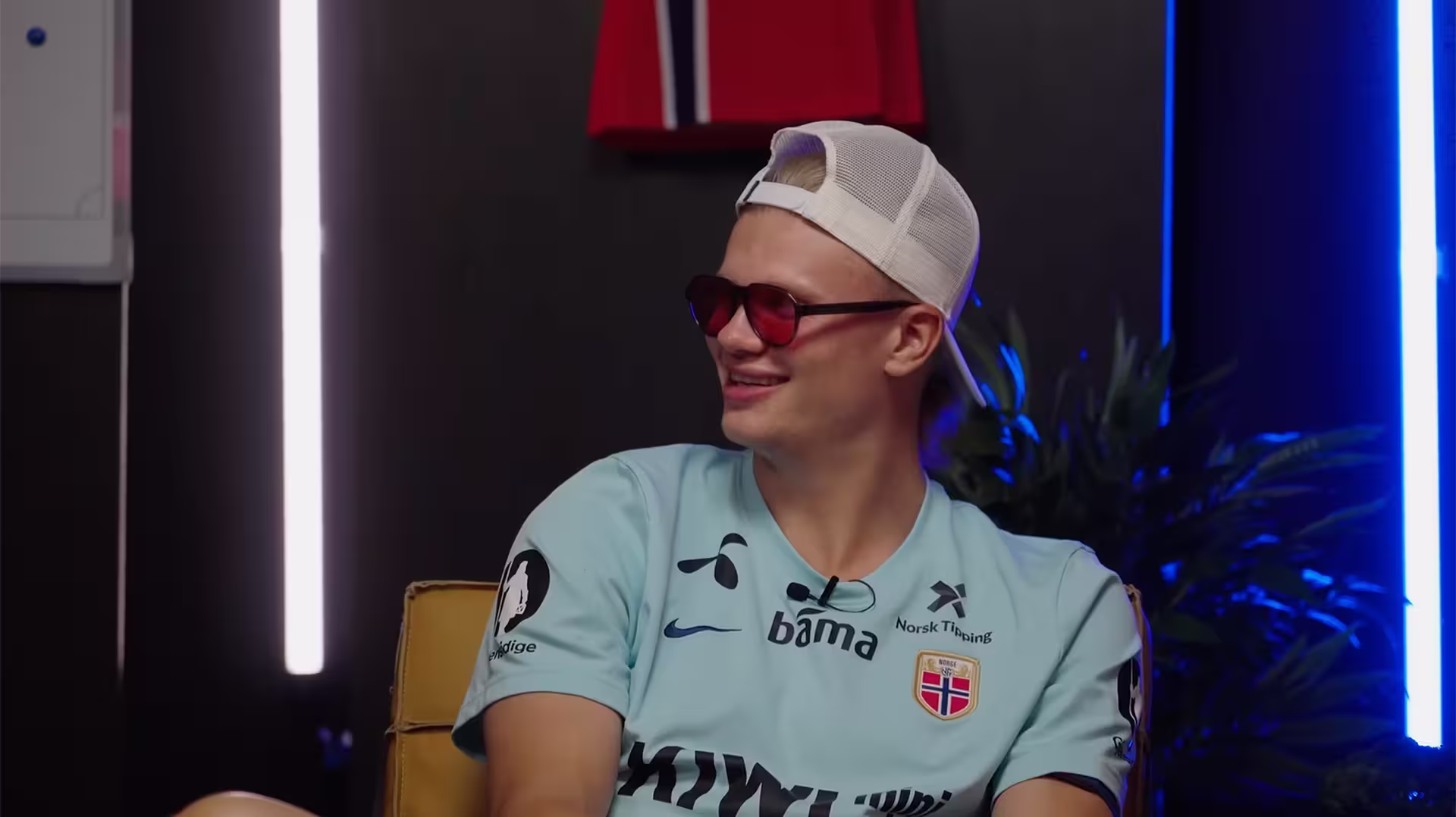 Young man in a light blue sports jersey, red sunglasses, and white cap turned backward smiling indoors.