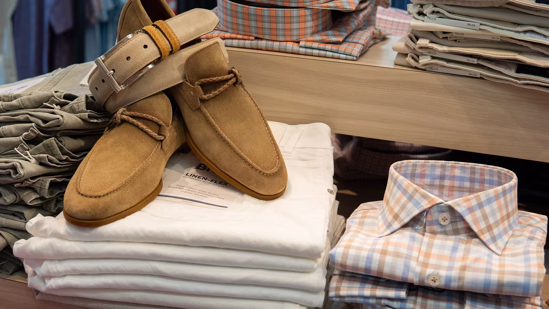 Tan suede loafers with beige belt atop white folded pants, next to folded plaid shirts on shelves.