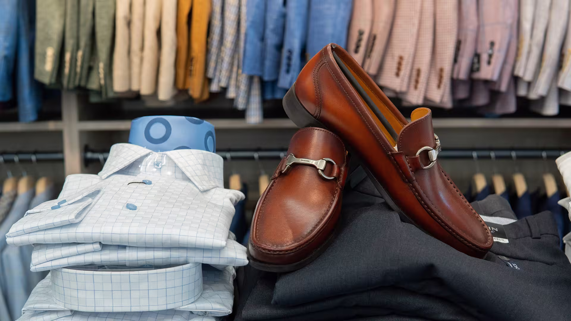 Brown leather loafers displayed on dark folded clothes beside stacked checked dress shirts in a men's clothing store.