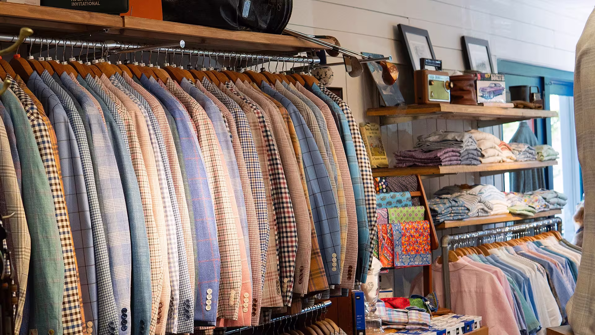 Tailored jackets and dress shirts hanging on racks in a men's clothing store with shelves of folded clothes.