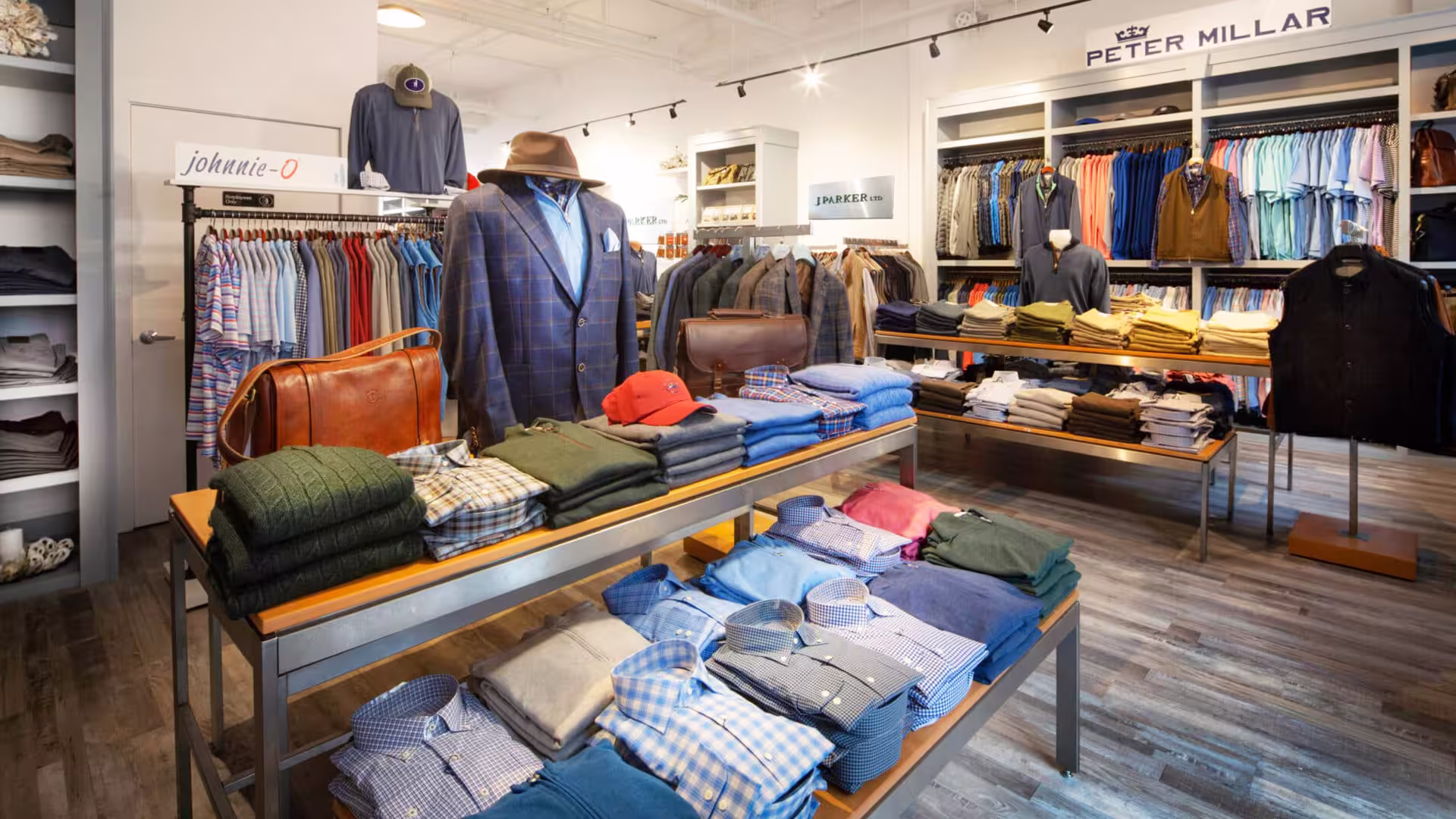 Men's clothing store with folded shirts, sweaters, jackets, hats, and leather bags on display tables and racks.
