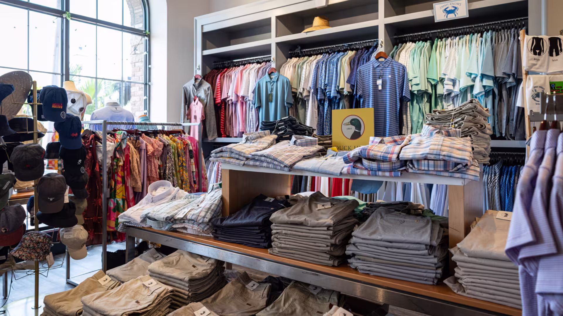 Clothing store interior with folded shirts, pants, hanging shirts, hats, and dresses on racks.