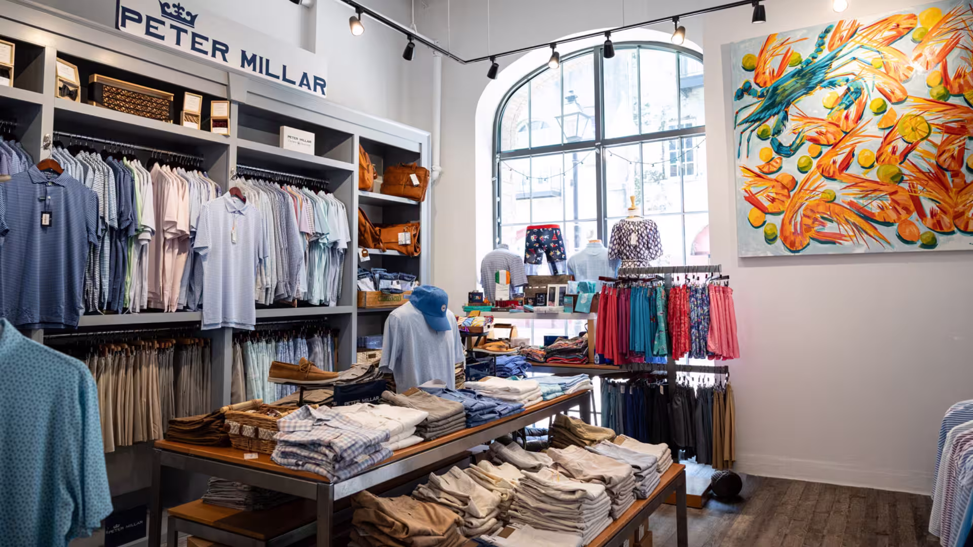 Clothing store interior with folded pants, shirts on racks, bright window, and colorful painting on wall.