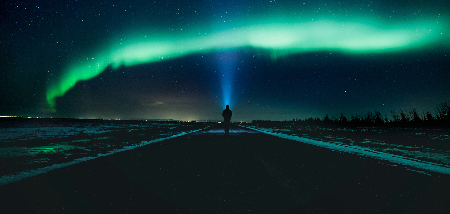 Person standing on a snow-covered road under a glowing green aurora borealis