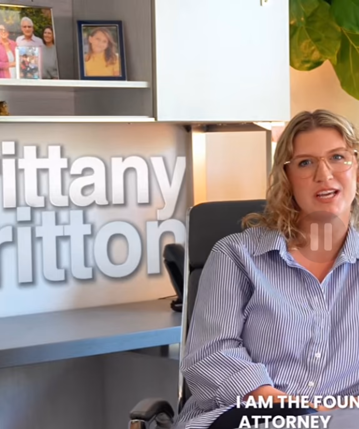 Woman with blonde curly hair and glasses speaking while seated in an office chair wearing a blue and white striped shirt; behind her is a shelf with framed photos and a large sign partially reading 'ittany' and 'ritton'.
