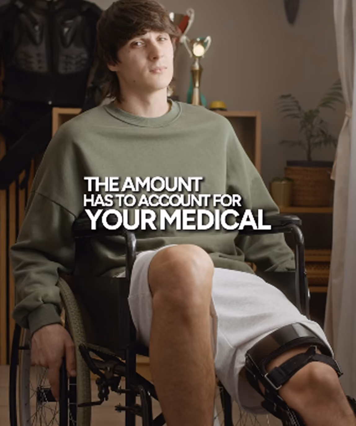 Young man in a wheelchair wearing a green sweatshirt and white shorts with a knee brace on his right leg, seated indoors with shelves and trophies in the background, with overlaid text reading 'THE AMOUNT HAS TO ACCOUNT FOR YOUR MEDICAL'.