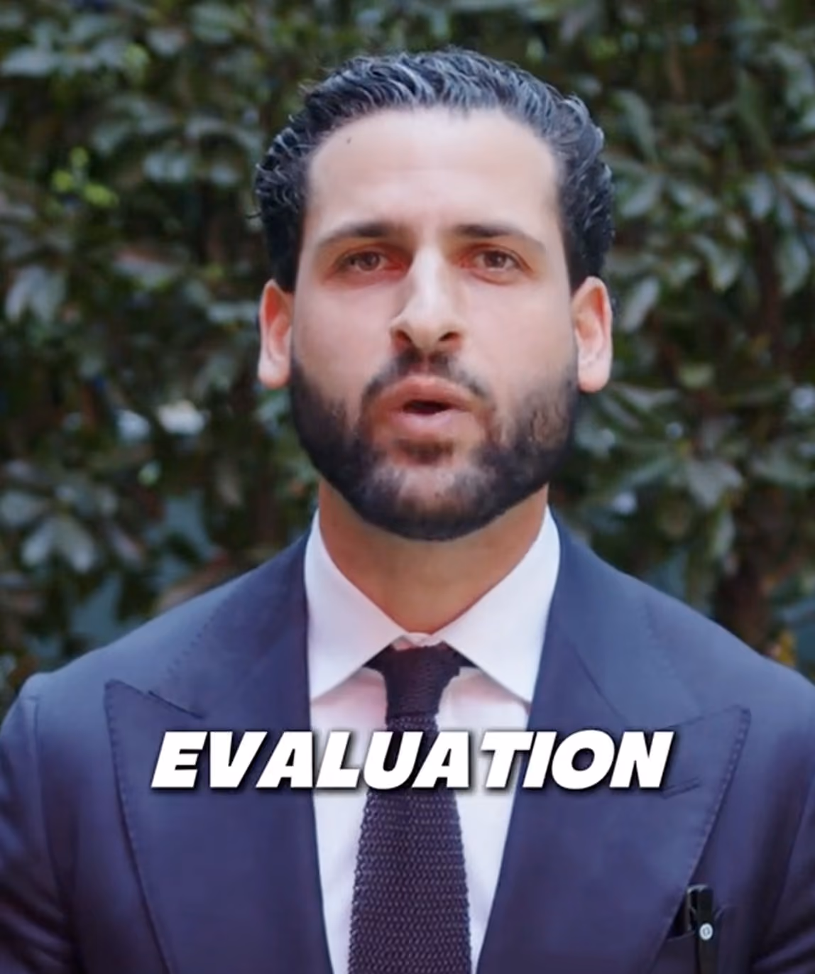 Man with dark hair and beard wearing a navy suit, white shirt, and dark tie standing outdoors with greenery background, with the word 'EVALUATION' in bold white text across his chest.