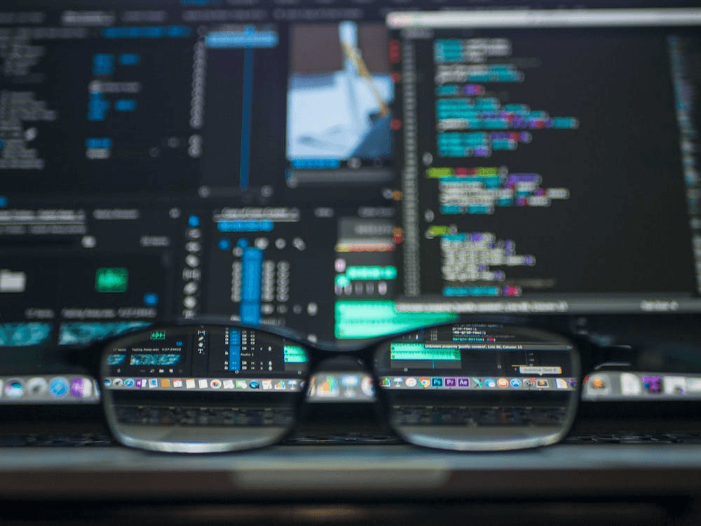 Decorative photograph of a pair of glasses in front of a monitor filled with colour-highlighted code.