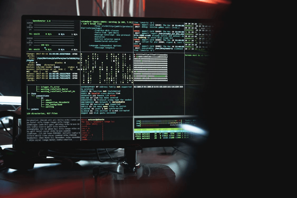 Decorative photograph of a darkened room where two monitors display dense scrolling terminal output in green and red.