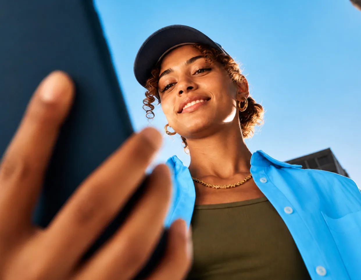 Joven sonriente con gorra azul mirando un teléfono móvil bajo un cielo despejado.