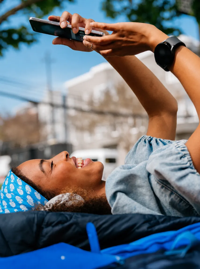 Smiling woman lying outdoors using a mobile phone and wearing a blue headband with white flowers.
