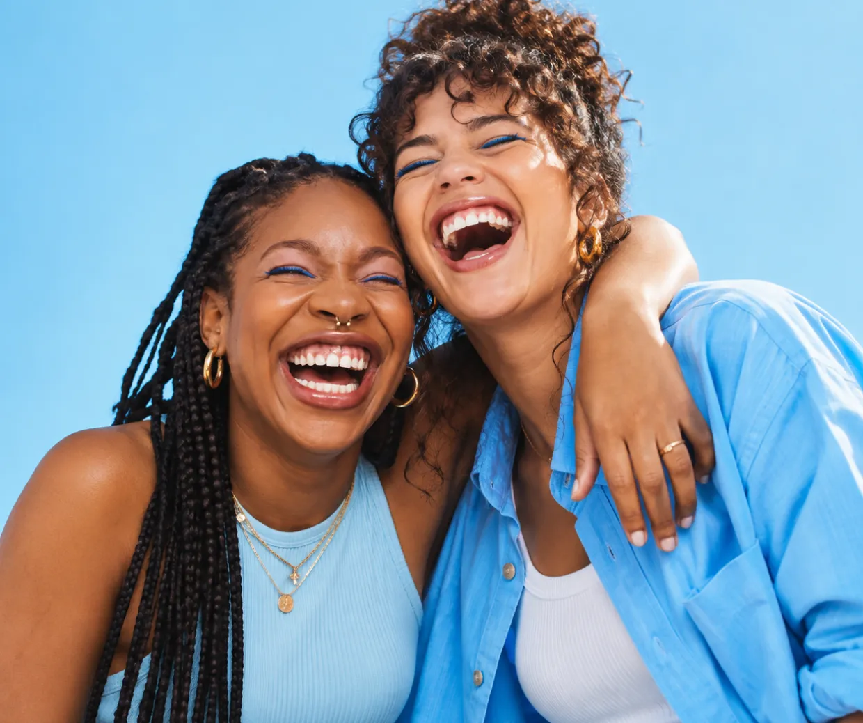Dos mujeres jóvenes sonrientes con maquillaje azul, una con trenzas y un piercing en la nariz, la otra con cabello rizado y camisa azul, abrazándose y riendo contra un fondo azul claro.