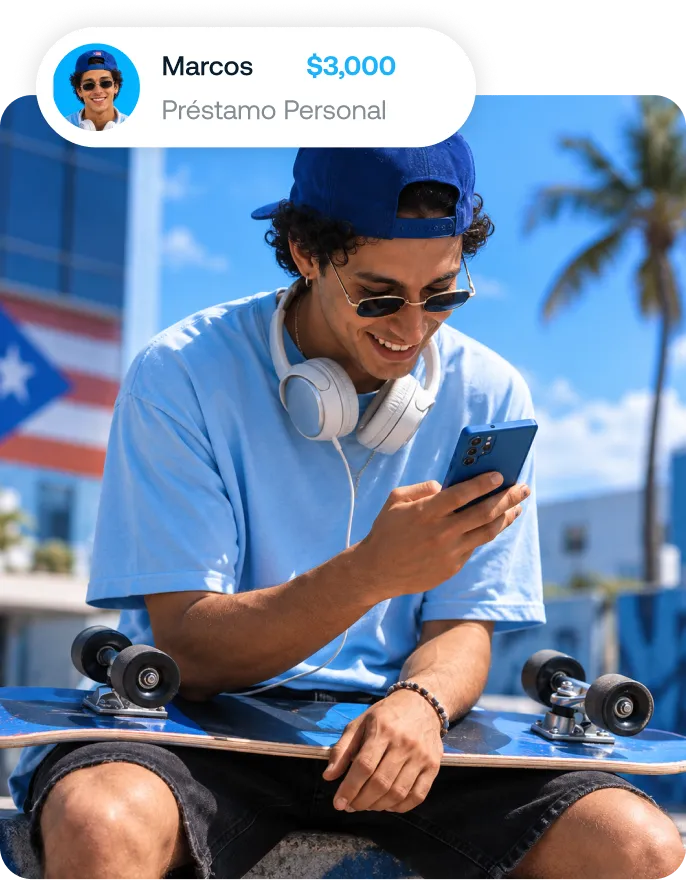 Joven con gorra azul, camiseta azul y auriculares blancos alrededor del cuello, sentado con un patineta en las piernas mientras sonríe mirando su celular azul al aire libre con edificio y palmera de fondo.