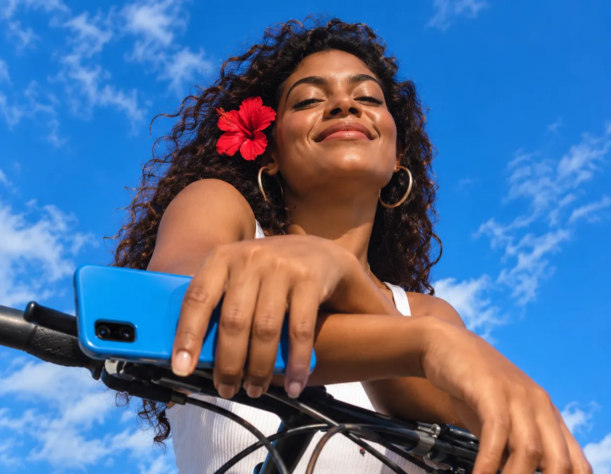 Mujer sonriente con flor roja en el cabello, usando aretes grandes, apoyada sobre el manillar de una bicicleta, con cielo azul y nubes blancas de fondo.