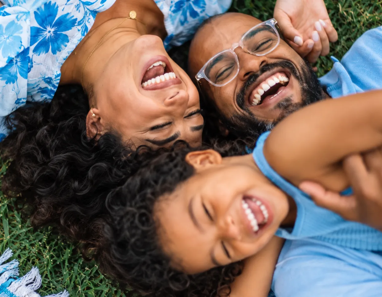 Familia con tres miembros acostados en el césped, riendo y disfrutando juntos, con prendas azules y cabello rizado.