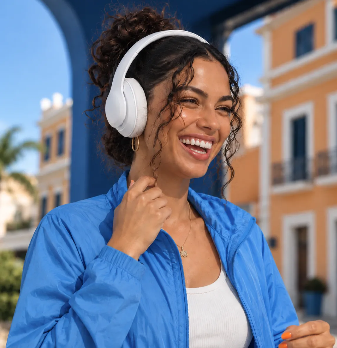 Mujer sonriente con audífonos blancos y chaqueta azul frente a edificios coloridos bajo cielo azul.