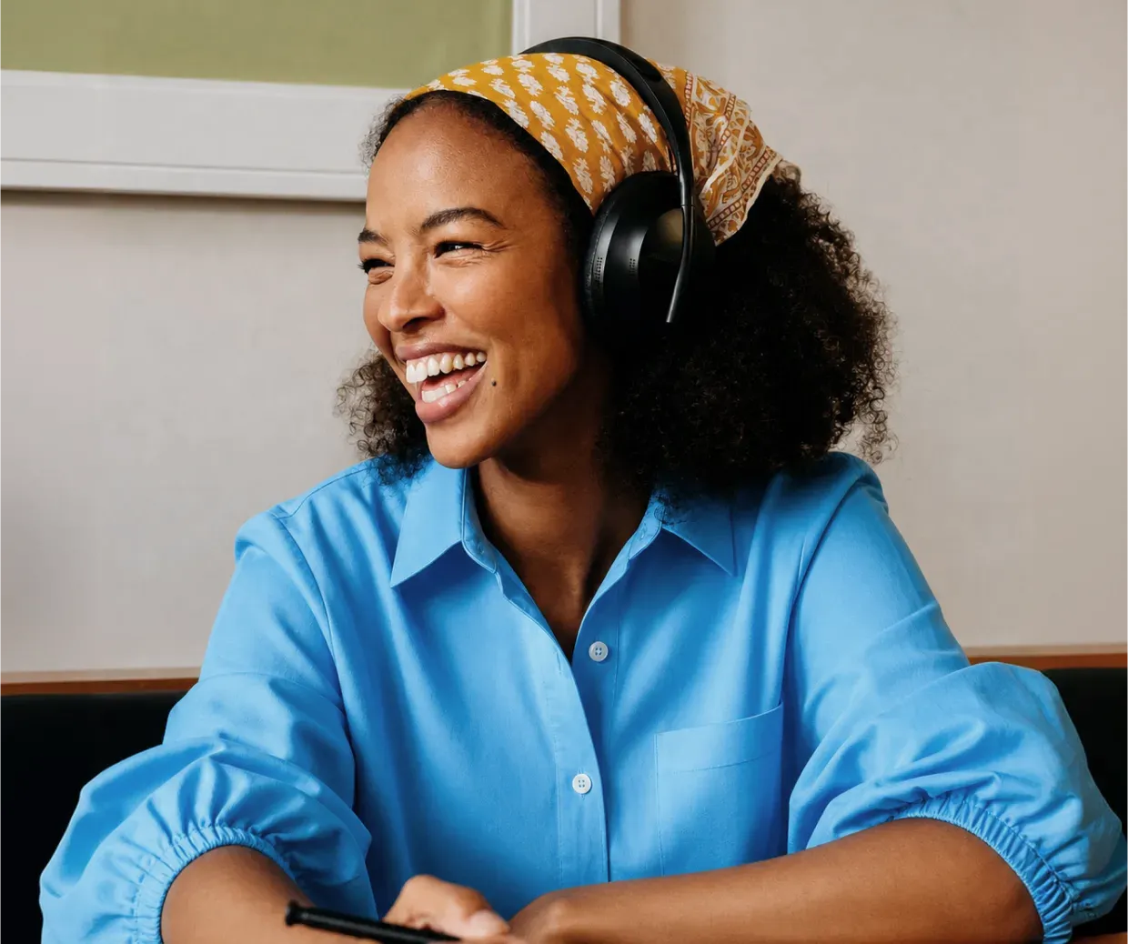 Mujer sonriente con auriculares negros y pañuelo amarillo en la cabeza, usando camisa azul.