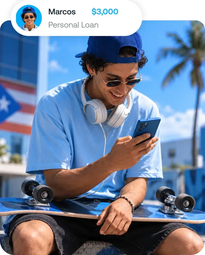 Young man in a blue cap, blue T-shirt and white headphones around his neck, sitting with a skateboard on his lap and smiling at his blue phone outdoors, with a building and a palm tree in the background.