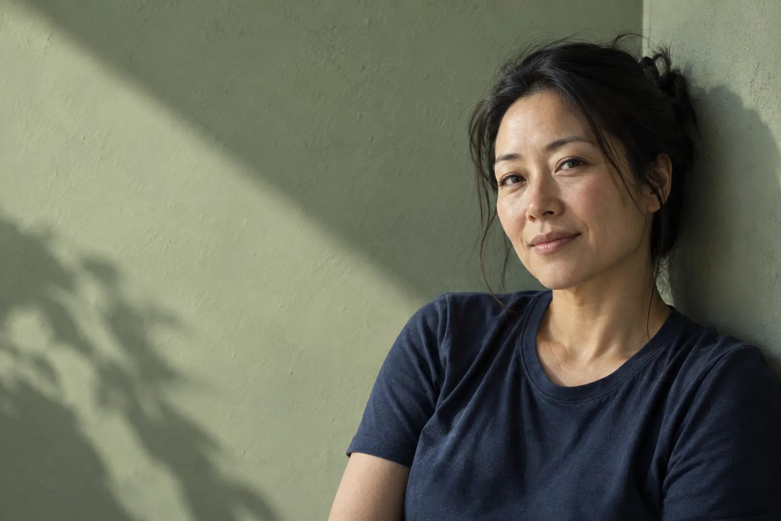 Woman in a navy cotton shirt resting beside a sage wall in sunlight