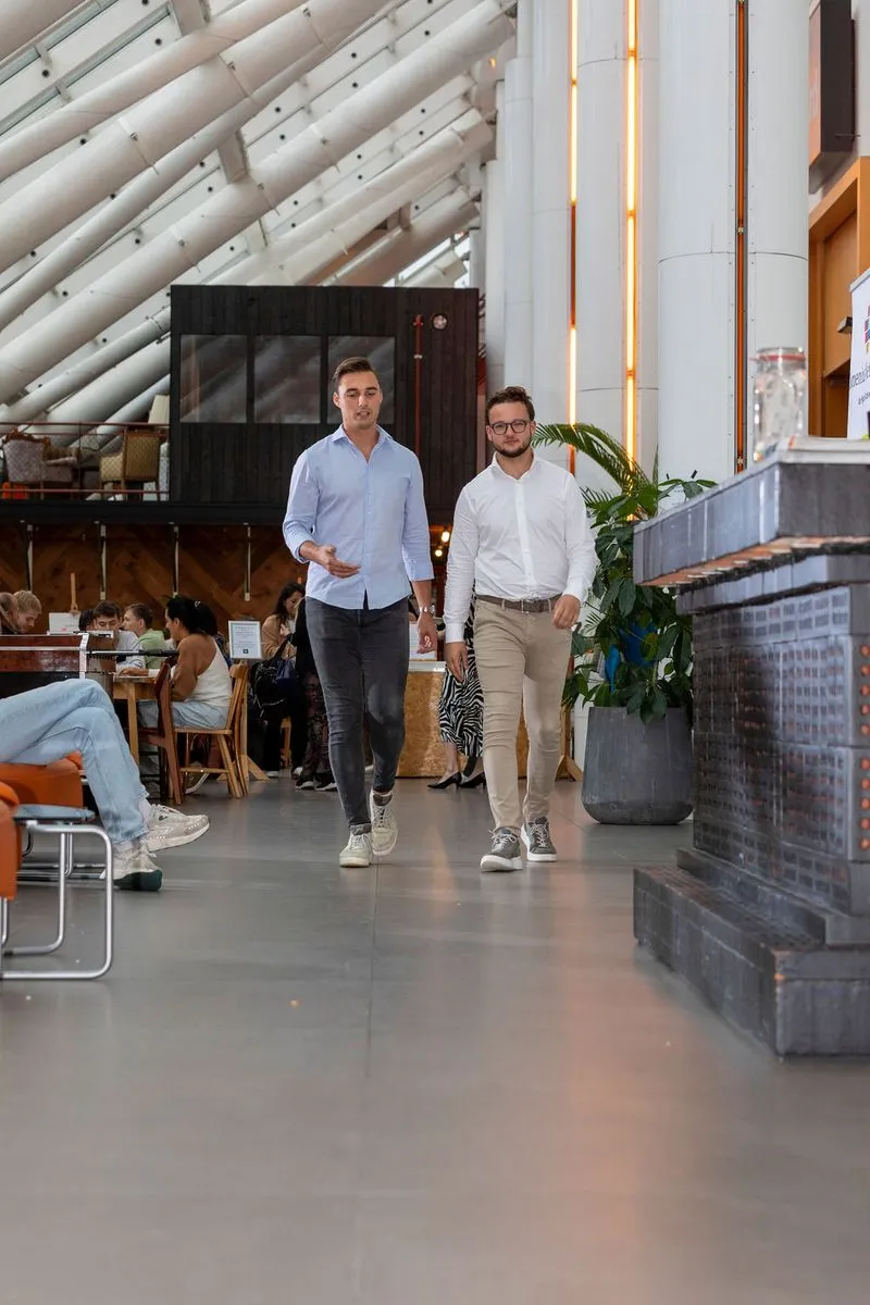 Twee mannen lopen en praten in een modern gebouw met mensen die aan tafels zitten.