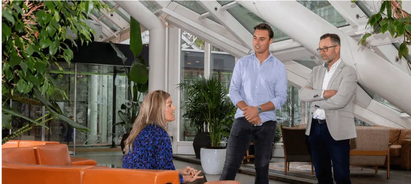 Drie personen in een modern kantoor met planten en oranje zitbanken, een vrouw zittend, twee mannen staand.