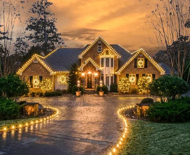 Professionally decorated suburban home at dusk with warm Christmas lights outlining the roof, windows, trees, and driveway.