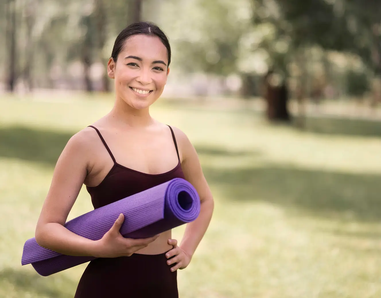 Smiling woman in workout clothes holding a purple yoga mat outdoors in a sunny park.
