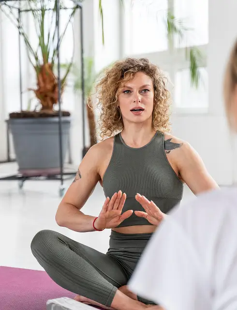 Woman with curly hair in gray workout clothes sitting cross-legged on a yoga mat gesturing hands.