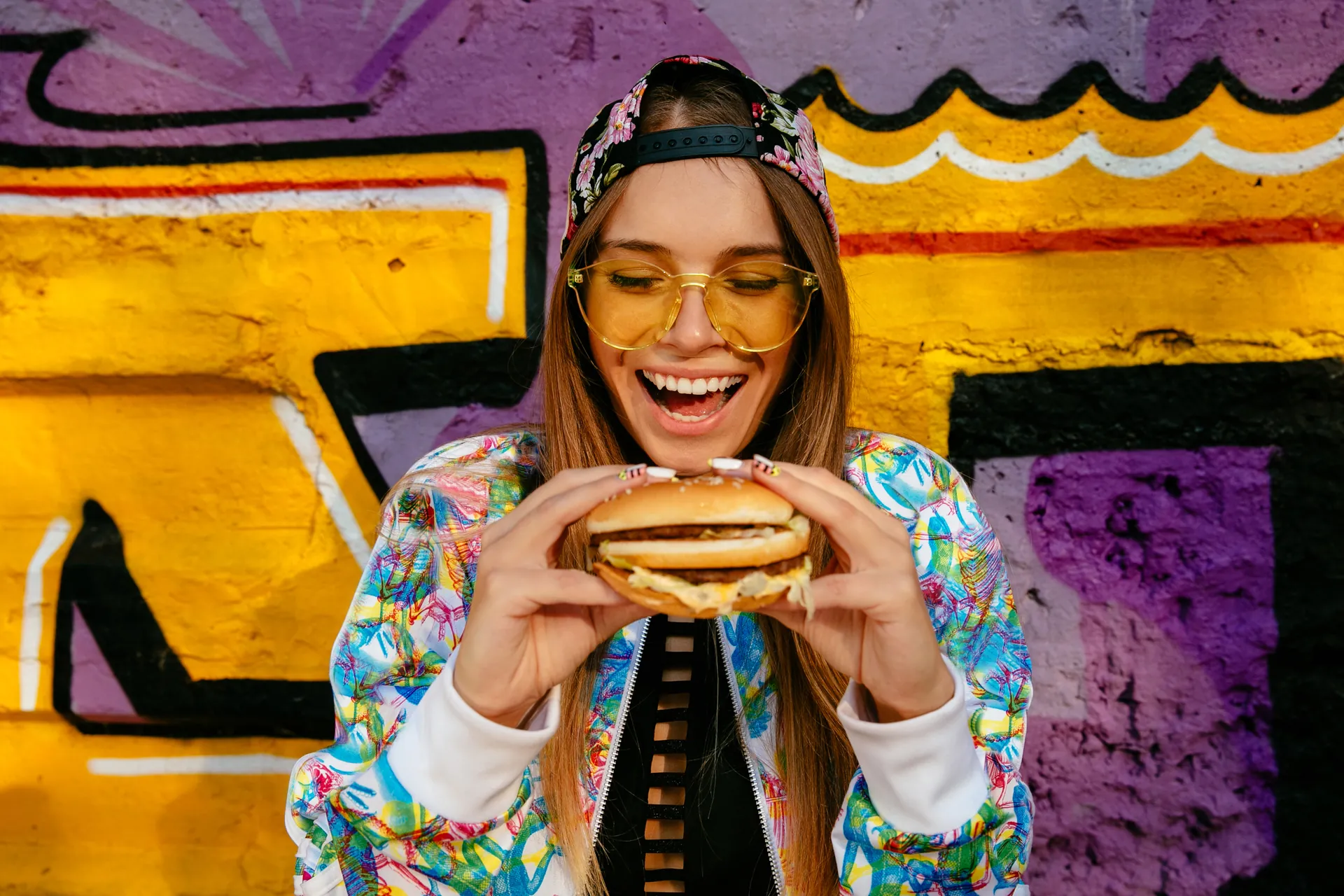 Junge Frau mit Sonnenbrille und bunter Jacke hält lachend einen Burger vor einer farbenfrohen Graffiti-Wand