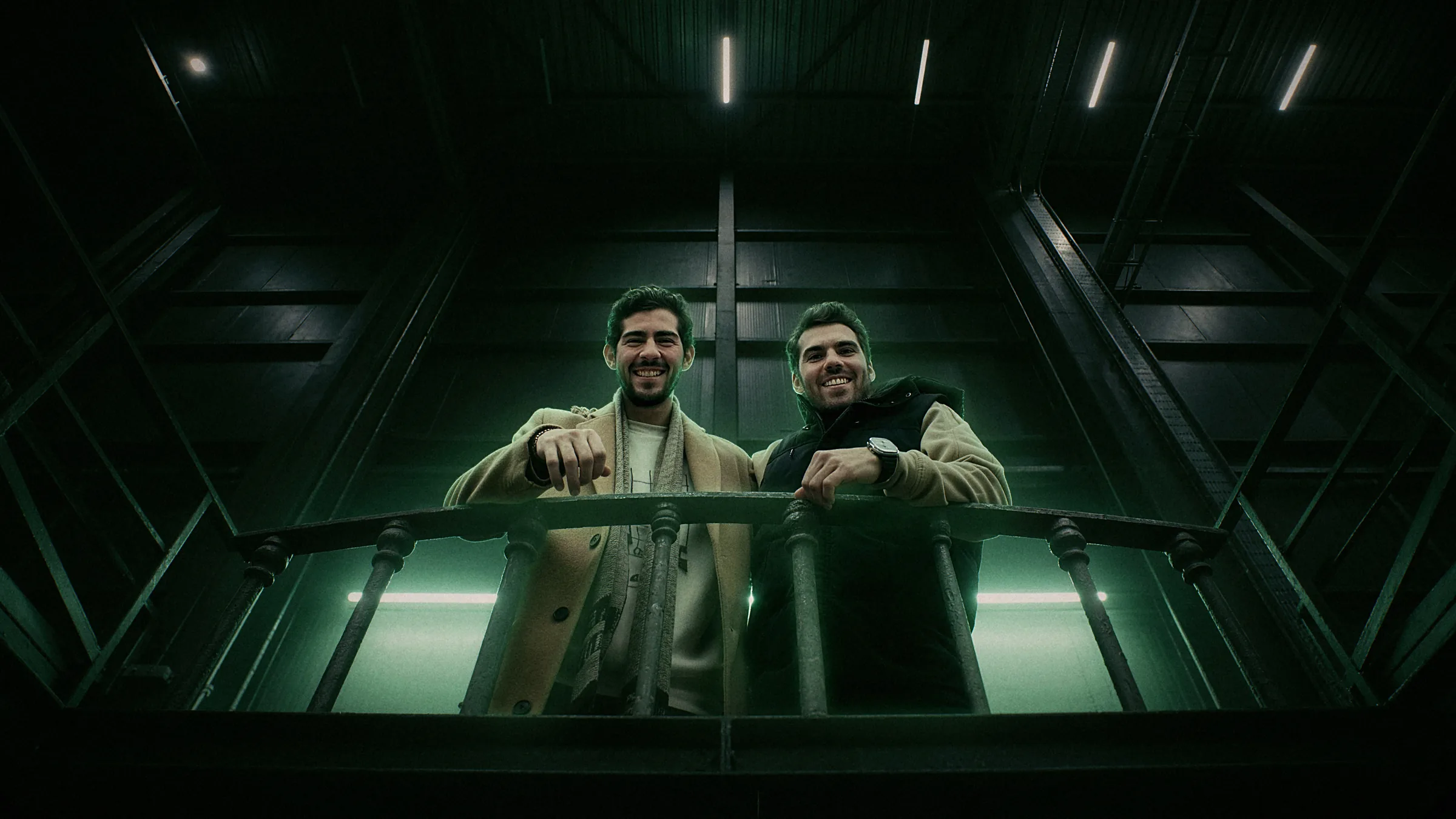 Andrés and Alejandro, Flamel's founders, leaning on a catwalk railing inside a dark factory hall.