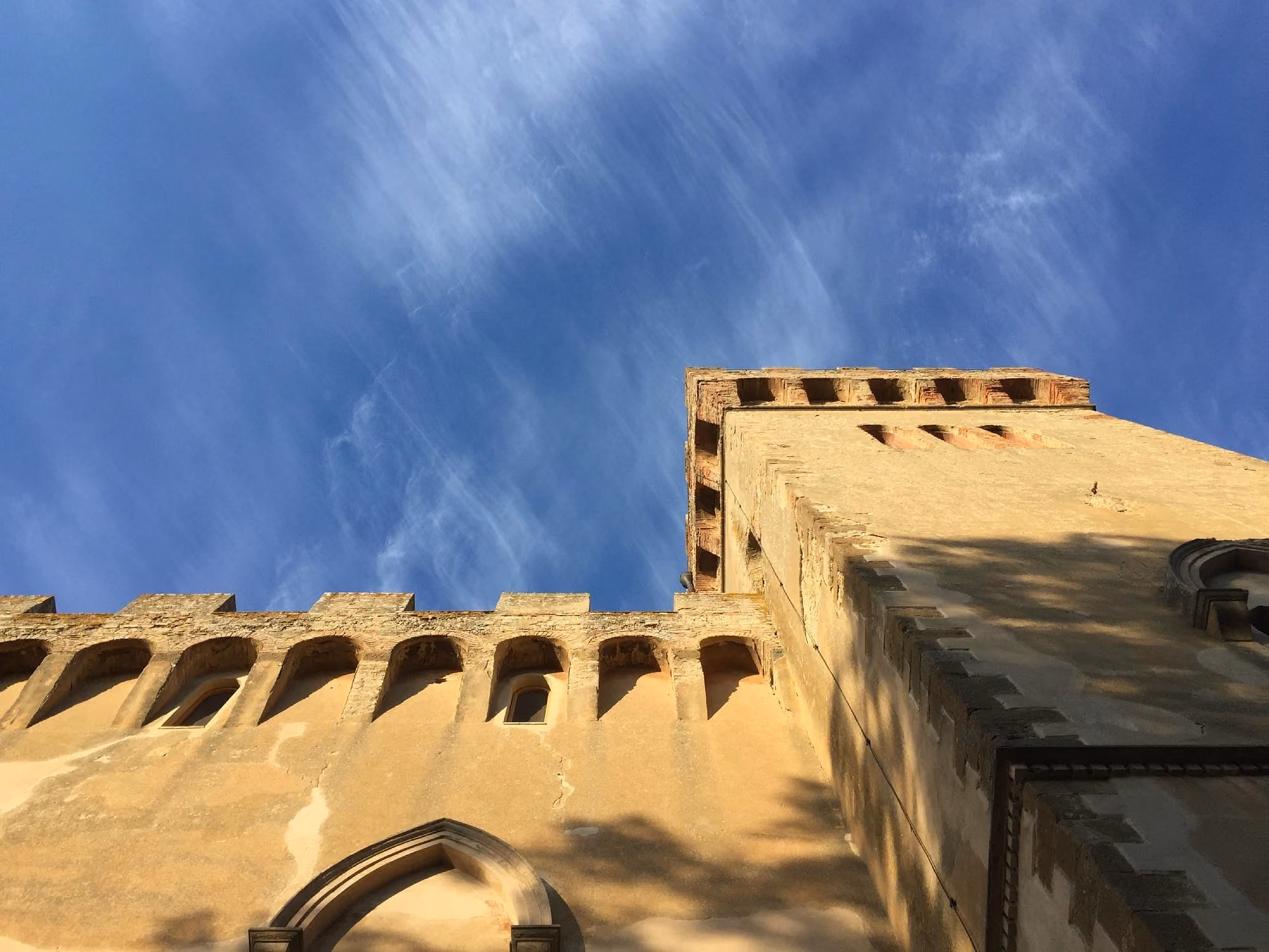 Low angle view of an old stone castle wall and tower with battlements under a blue sky with wispy clouds.